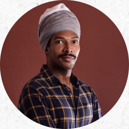 Foto de um homem negro, olhos escuros e bigode escuro que posa para a camêra. Ele usa uma camisa xadrez com fundo azul escuro e linhas alaranjadas. Ele usa um turbante de tecido cinza com uma parte branca no topo. O fundo da imagem é marrom. 