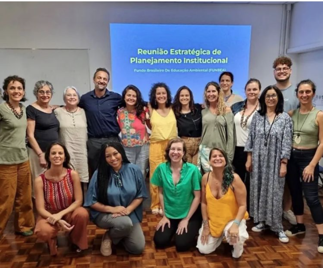 Foto de um grupo de 17 pessoas, a maioria mulheres brancas de meia idade. Todos usam roupas coloridas e posam para a foto, alguns em pré, abraçados, e outros agachados à frente. O chão é de tacos de madeira e ao fundo há uma parede branca com a frase “reunião Estratégica de Planejamento Institucional: Fundos Brasileiros De Educação Ambiental (FunBEA)” projetada.