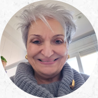Na foto, uma mulher branca, idosa, de cabelos brancos e curtos sorri. Ela usa brincos dourados e uma blusa de lã cinza.