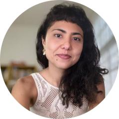 Foto de mulher com traços amarelos, olhos escuros e cabelos longos, ondulados e escuros. Ela tem uma pinta abaixo dos lábios e veste uma camiseta regata branca. 