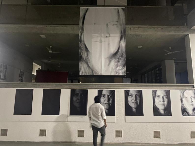 Foto de um homem observando uma exposição de fotografias. Ele tem cabelos curtos e escuros, veste uma camisa branca e uma calça cinza e está de costas. A exposição é formada por oito imagens. Sete delas estão fixadas lado a lado em um muro branco. A oitava imagem é maior que as outras e está suspensa, acima do muro. As fotografias são do rosto de uma mulher, que encara seriamente a câmera. As imagens posicionadas à esquerda são mais escuras, e passam a ser mais claras e iluminadas conforme estão à direita.