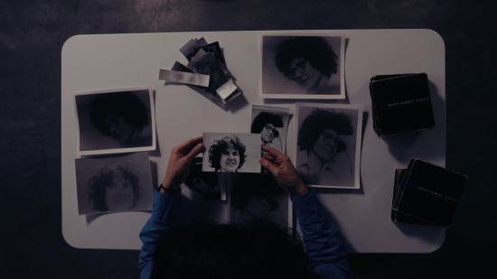 Foto, a partir do alto, de uma pessoa segurando e observando uma fotografia preto e branco em uma mesa retangular branca sobre a qual estão outras fotografias preto e branco, mais escuras. A pessoa tem cabelos escuros, veste camisa de mangas longas azuis e tem a pele clara. Com as duas mãos, ela segura  uma foto do rosto de Norma Bahia, uma pessoa de pele clara e cabelos curtos escuros.