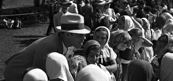 Na fotografia em preto e branco, Mário de Andrade, um homem branco com cabelos castanhos. Ele usa um chapéu claro com uma faixa preta, óculos e um terno escuro. Ele está abaixado e conversa com várias crianças que têm véus claros sobre a cabeça. Ao fundo, homens e mulheres conversam entre si.