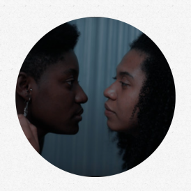 Foto de uma cena do curta. Duas mulheres negras se olham profundamente nos olhos. A da esquerda usa um brinco e tem cabelo crespo e curto. A da direita tem cabelo cacheado e volumoso. Ela coloca sua mão esquerda no pescoço da outra mulher.
