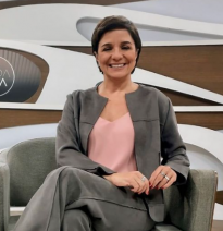 Foto de uma mulher branca sentada sorrindo. Ela tem cabelos lisos, curtos e escuros. Sua roupas são rosa claro e cinza. Nas suas mãos, que estão apoiadas no colo, ela usa alguns anéis. A poltrona em que está sentada é cinza. O fundo é abstrato, nas cores marrom e branco.