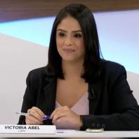 Foto de uma mulher sentada segurando uma caneta. Ela é branca, tem cabelos lisos e escuros e olhos também escuros. A mulher veste uma blusa clara e um blazer preto. Ela apoia suas mãos em uma bancada, onde também estão outros objetos, como um celular e uma placa com seu nome e empresa: Victoria Abel CBN. Ela sorri levemente e não olha diretamente para a câmera.