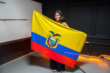Foto de uma mulher de pé, sorrindo e segurando, à frente de seu corpo, com os braços abertos e inclinados diagonalmente, a bandeira do Equador. A mulher tem a pele morena e cabelos lisos e escuros de tamanho médio..