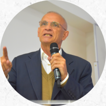 um homem branco, com cabelos ralos e grisalhos, fala segurando um microfone na mão. Com a outra, ele aponta para frente com o dedo indicador. Ele usa oculos e veste uma camisa branca, um sueter bege e um paletó azul.