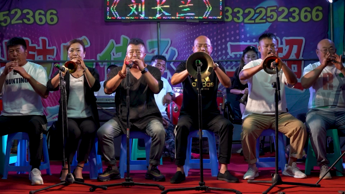 Na foto, seis pessoas de origem chinesa estão sentadas em bancos de plástico brancos, tocando instrumentos de sopro. À frente de cada uma delas, há um tripé que segura um microfone. O grupo é composto por pessoas de diferentes gêneros que vestem roupas casuais, a maioria em tons de branco e preto. O chão está coberto por um carpete vermelho, e, ao fundo, há um painel colorido com números e palavras em chinês.