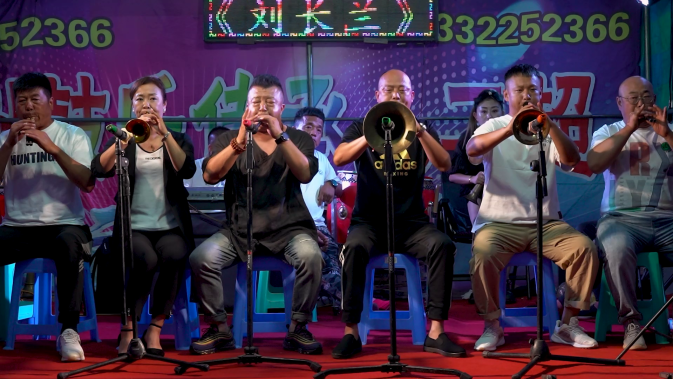 Na foto, seis pessoas de origem chinesa estão sentadas em bancos de plástico brancos, tocando instrumentos de sopro. À frente de cada uma delas,  há um tripé que segura um microfone. O grupo é composto por pessoas de diferentes gêneros que vestem roupas casuais, a maioria em tons de branco e preto. O chão está coberto por um carpete vermelho, e, ao fundo, há um painel colorido com números e palavras em chinês.