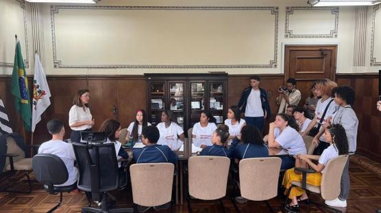 Foto de uma coletiva de imprensa do projeto Memórias em Rede. Há cerca de 18 adolescentes. 14 estão sentados em volta de uma mesa ovalada e 4 deles estão de pé. A maioria usa camisetas brancas e alguns usam agasalho azul escuro. À esquerda da imagem, há uma mulher branca de pé, de cabelos castanhos claros curtos, óculos e camisa branca. Estão em uma sala com piso de madeira e parede beges, com bandeiras do Brasil, do Estado de São Paulo e da cidade de Santos. 