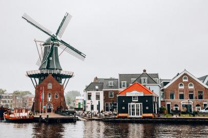 Foto colorida em ambiente externo, de um moinho de vento holandês às margens de um porto, com um céu nublado ao fundo