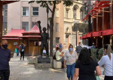 Foto da estátua de Zumbi dos Palmares. O retratado é negro, está descalço e usa apenas uma calça. Ele tem o braço direito erguido, com o punho cerrado, e na mão esquerda tem uma lança apoiada paralelamente ao corpo, com umas das extremidades tocando o chão. A obra está ao lado de uma árvore e cercada de quiosques com tendas vermelhas. Na praça, diversas pessoas circulam, usando máscaras. 