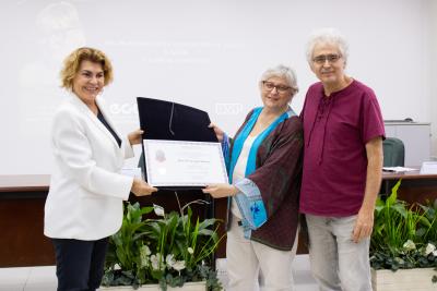 Três pessoas posam de pé para uma fotografia. Duas mulheres seguram uma pasta preta aberta, uma de cada lado, exibindo um certificado. À esquerda, está uma mulher loira, de cabelos curtos, vestindo um blazer branco, camisa e calças pretas. Do outro lado, está uma mulher branca, de cabelos brancos, lisos e curtos. Ela usa óculos de haste avermelhada, veste calça e blusa brancas e uma blusa, do tipo quimono, vermelho escuro com detalhes em azul claro. Do lado direito da imagem, abraçando a segunda mulher, est