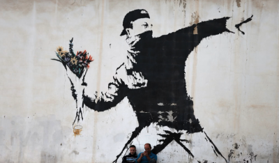 Foto de grafite na cor preta sobre um muro claro. O desenho retrata um homem com boné e rosto coberto segurando um buquê de flores, em um gesto de quem se prepara para lançar o objeto, semelhante ao gesto de quem lança uma granada ou um coquetel Molotov. As flores são amarelas e vermelhas.