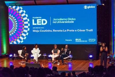 Foto de cinco pessoas em um palco diante de uma plateia. Quatro delas, duas mulheres negras, uma mulher branca e um homem, estão sentadas em poltronas brancas. O homem fala ao microfone e gesticula olhando para a plateia, enquanto as mulheres o olham. No canto direito do palco, uma mulher negra está de pé fazendo linguagem de sinais. No fundo do palco, um grande telão exibe os dizeres ‘Academia LED. Jornalismo Globo na Universidade. Aula aberta com Maju Coutinho, Renata Lo Prete e César Tralli. 
