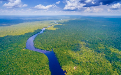 Foto de floresta vista de cima, cortada por um rio que segue do primeiro plano até o fundo da imagem. O céu é azul com nuvens. 