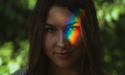 Foto de uma mulher de cabelos escuros e olhos claros. Sobre um de seus olhos, a projeção de um arco-íris. Ao fundo, folhagens desfocadas. 