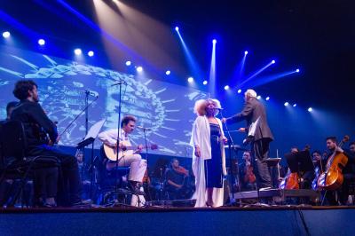 Foto de um palco durante um concerto. No centro, uma mulher negra, de cabelos crespos e loiros, está em pé diante de um microfone, usando um vestido escuro e um casaco branco e longo. Ao lado dela, um maestro de cabelos brancos usando um terno gesticula com uma das mãos erguidas, de frente para a orquestra. Atrás da cantora, um músico de roupas claras toca um violão sentado. À esquerda e à direita, membros da orquestra estão sentados com violinos, violoncelos e outros instrumentos.