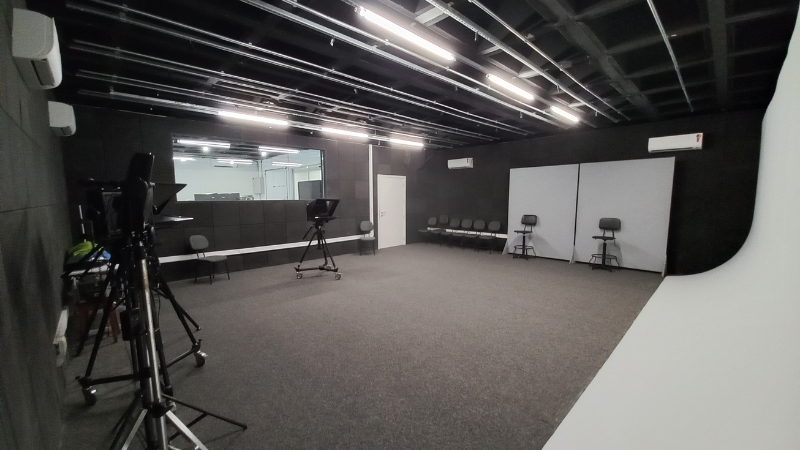 Estúdio de gravação de TV: todo em tapete na cor cinza, fundo infinito na cor branca, à direita e, à esquerda, janela de vidro. O estúdio tem ainda equipamentos de gravação - abril de 2022