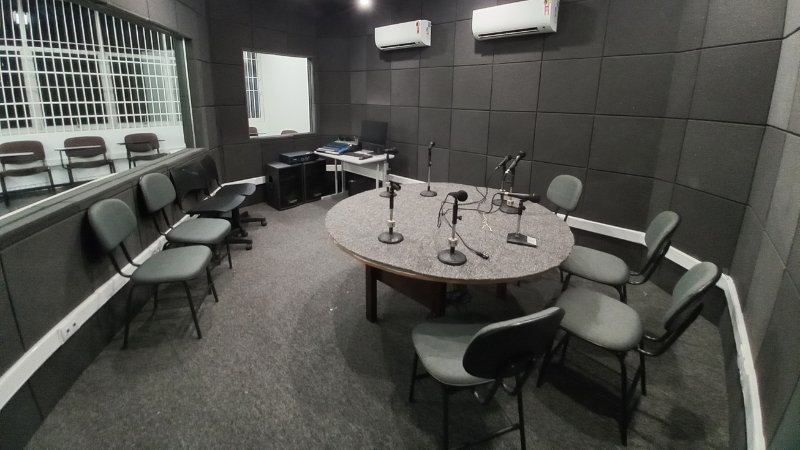 Sala de gravação do estúdio de rádio. Uma mesa redonda, com microfones em cima e cadeiras ao redor - abril de 2022 O estúdio tem tapete de tecido na cor cinza e peças para isolamento acústico, também na cor cinza.