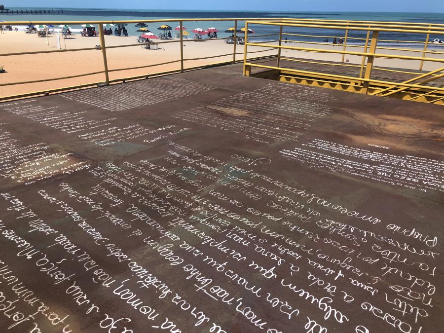 Resultado da intervenção no topo do contêiner. Diversas mulheres escreveram nele suas histórias com a praia de Iracema. Foto: Marie Auip. Foto de um piso marrom, cortado por uma escada e cercado por um guarda-corpo amarelo, com escritas brancas em diferentes caligrafias. Ao fundo,uma praia com areia alaranjada, mar azul e vários guarda-sóis coloridos.