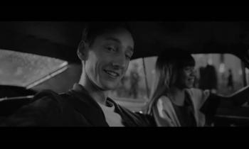 imagem em preto e branco de campanha publicitária em que aparecem um jovem branco, que sorri e olha para a camera, e ao fundo uma mulher, levemente desfocada, que também sorri enquanto olha a frente. eles estão dentro de um carro.