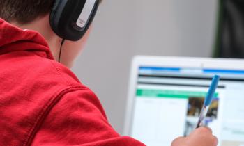 Foto de um garoto escrevendo, enquanto usa um laptop e fones de ouvido. O menino, que é branco e possui cabelo escuro, usa uma blusa de moletom vermelha e fones de ouvido pretos e cinzas. Ele segura com a mão direita uma caneta transparente de tampa azul. Ao fundo, há um laptop branco com a tela desfocada. Em último plano, está uma parede branca.