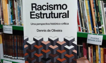 uma mão negra segura um livro, ao fundo, prateleiras com vários livros.