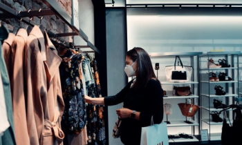 Foto de uma mulher escolhendo roupas em uma loja. A mulher é branca, possui cabelos lisos e escuros e está usando máscara. A roupa dela é preta e ela está com um relógio no braço esquerdo, no qual pendura uma sacola de compras. Essa mulher está escolhendo roupas entre várias opções penduradas em uma arara. Ao fundo, pode-se ver prateleiras com bolsas e sapatos.