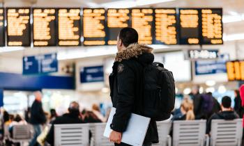 em um aeroporto, um homem está de pé, vestindo um caso preto e segurando um laptop na mão. Ele olha para um painel ao fundo, ligeiramente desfocado. à sua frente, também um pouco mais ao fundo, estão cadeiras brancas e pessoas sentadas de costas para ele.