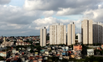 Foto de um bairro da região leste de Osasco. É possível observar uma comunidade composta por casas de baixo padrão cercada por prédios residenciais de classe média. O céu, ao fundo, tem muitas nuvens.