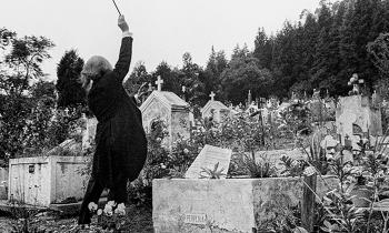 Fotografia em preto e branco. De costas para a câmera, um maestro de cabelos grisalhos na altura da nuca faz movimentos com a batuta, como se estivesse regendo uma orquestra, diante de lápides e mausoléus em um cemitério.