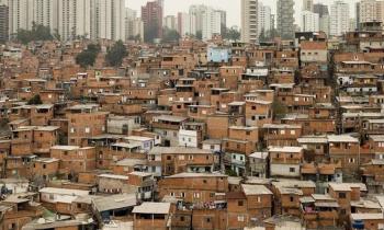 Foto de uma favela tirada do alto. Diversas casas estão bem próximas e não estão rebocadas. Bem ao fundo, estão alguns prédios ca