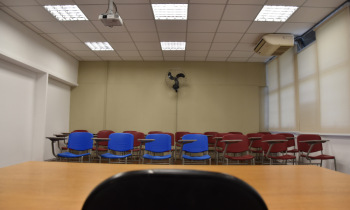 Foto de uma sala de aula. Em primeiro plano está uma mesa de madeira, de onde a foto foi tirada, e uma cadeira. Ao fundo, estão mais cadeiras, que são vermelha e azuis. A parede do fundo é bege e há um ventilador nela.