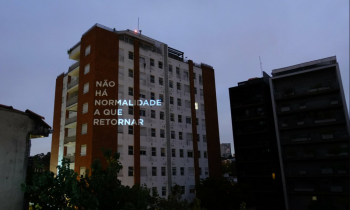 foto de um fim de tarde nublado, de um prédio branco e marrom. Nele, está projetada em luz a frase não há normalidade a que retornar.