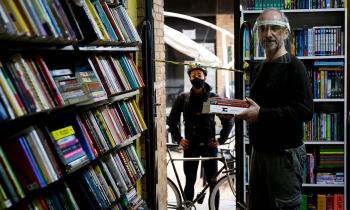 a esquerda, uma estande chei de livros; à direita, um homem branco e calvo, veste roupas pretas e um face shield, segura dois livros com as mãos. Ao fundo, outro homem branco, vestido de preto, posa com as mãos na cintura, à frente dele, uma bicicleta.