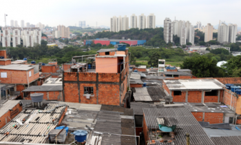 Foto tirada do alto apresentando casas populares em primeiro plano, com muitas paredes sem reboco. Ao fundo, algumas árvores e prédios distantes.