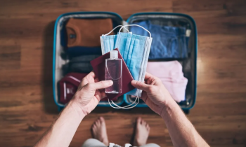 ao centro da imagem, duas mãos seguram duas máscaras, um frasco de alcool em gel e um passaporte. Ao fundo, uma mala aberta com roupas dobradas