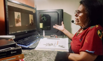 foto de uma senhora 0 branca, de cabelos de tamanho médio e levemente ondulados, oculos, que veste uma blusa vermelha. Ela olha para um monitor de computador, em que conversar via chamada de vídeo com outras duas pessoas. Ela está sentada em uma escrivaninha e, à frente dela, há um livro aberto.