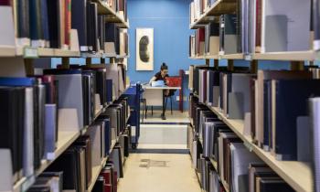 Foto tirada em um corredor formado por prateleiras de livros. Ao fundo, na frente de uma parede azul, uma jovem está sentada em uma mesa. Ela é branca, tem cabelos lisos escuros amarrados, usa óculos e veste preto. 