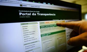 Foto da tela de um computador. Na tela, o site do Portal da Transparência do governo federal. O site possui colunas em cores verde claro e escuro. No canto direito da imagem aparece uma mão com o dedo indicador apontando para a tela. 