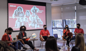 Foto de seis diretores da ECA Jr., homens e mulheres, sentados em frente à projeção com uma foto da equipe gestora. Eles vestem a camiseta vermelha da empresa.  No canto inferior da imagem, alguns estudantes assistem a apresentação.  Todos usam máscaras.