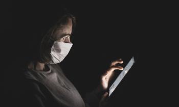 Foto mostra uma mulher branca de meia-idade com cabelos na altura dos ombro e olhos castanhos. Dentro de uma sala escura, ela usa máscaras e manuseia um tablet que ilumina seu rosto.