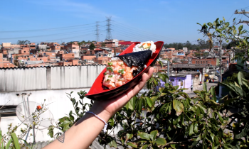 Foto mostra a mão de uma pessoa que segura uma barca de sushi contendo um temaki de salmão e um sushi. Ao fundo, o pedaço da copa de uma árvore e a vista de uma comunidade, com diversas casas de tijolo aparente. No horizonte há linhas de transmissão elétrica.