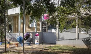 Foto da fachada do prédio do CMU, que tem cores claras. Na entrada, há uma escada e uma rampa. Na frente do prédio, além de muitas árvores, há alguns bancos, bicicletas e telefones “orelhões”. A placa do prédio tem um tom rosa forte e a sigla CMU em letras brancas.