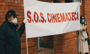 Foto de duas pessoas em frente à Cinemateca. Elas seguram uma faixa branca com a escrita vermelha: "S.O.S. CINEMATECA".