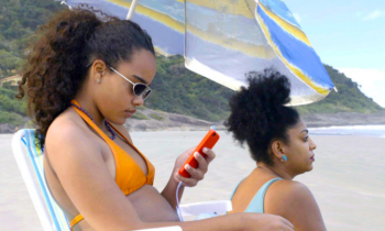 Imagem do filme "Receita de Caranguejo". Há, na foto, duas mulheres pretas em uma praia. A mulher em primeiro plano, sentada e usando óculos e biquíni amarelo, olha para seu celular de capa laranja. A outra, de biquíni e brinco azul, olha para a paisagem. Ao fundo, pode-se observar uma montanha e parte do mar.