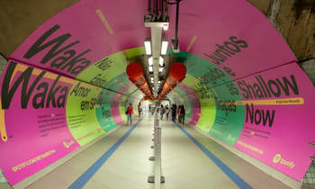 Foto tirada na ponta de um corredor no metrô de São Paulo . As paredes estão totalmente cobertas por cartazes publicitários chamativos (predominantemente em cor-de-rosa e em dois tons de verde) com títulos de músicas famosas escritas em preto. O corredor é bem iluminado. Nele, algumas barras de segurança separam as pessoas que andam nos dois sentidos.