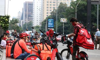 Foto de entregadores de aplicativos parados com suas bags (bolsas térmicas) e bicicletas aguardando pedidos na região da Avenida Paulista, em São Paulo. Suas roupas e capacetes são vermelhas, assim como as bags e bicicletas. Alguns têm máscaras abaixadas, encostando no queixo. Um deles está de pé checando o celular enquanto os demais permanecem sentados.